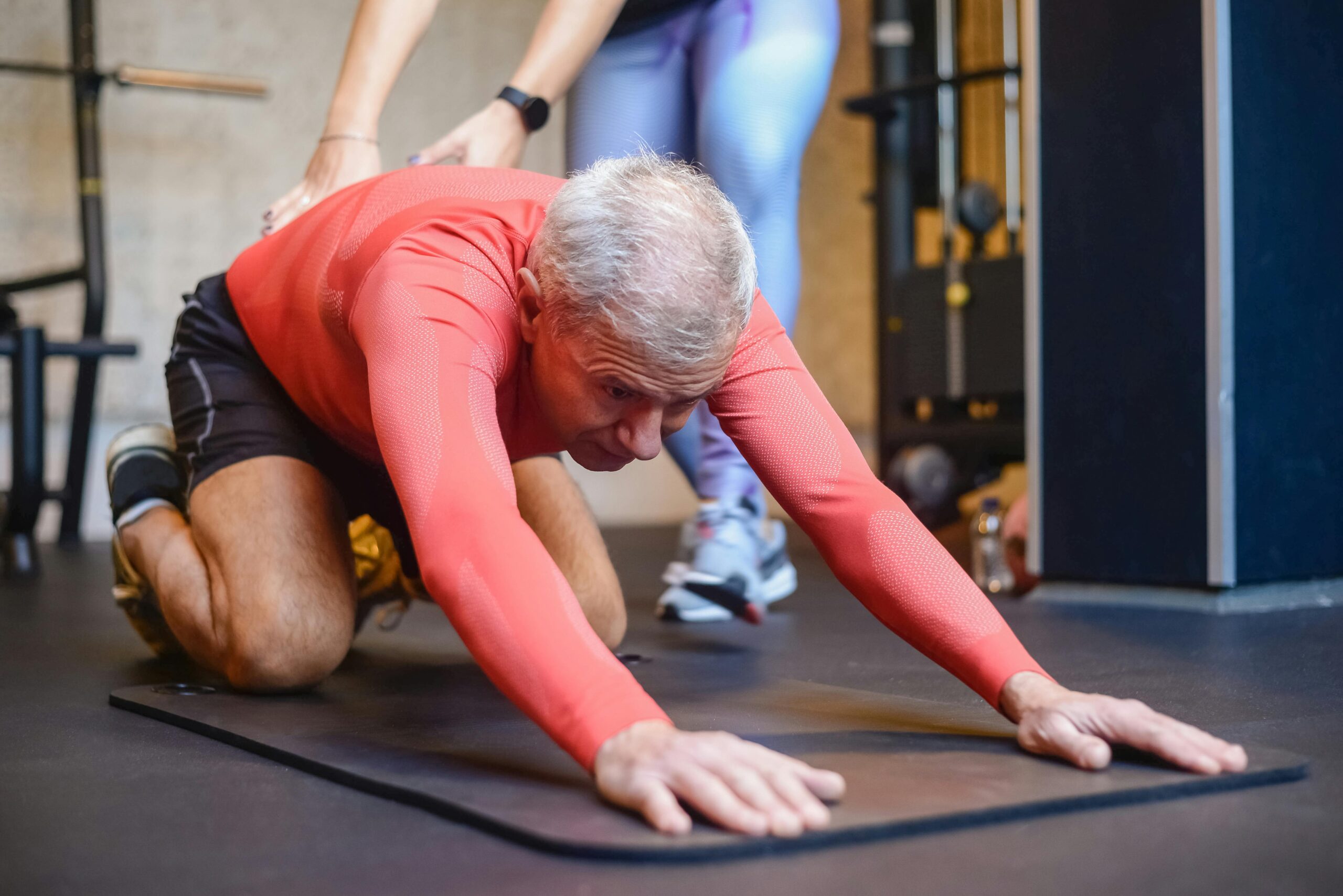 Senior man practicing yoga in a gym, stretching on a mat with the support of a trainer. He is wearing a red long-sleeve sports shirt and black shorts, focusing on flexibility and movement.