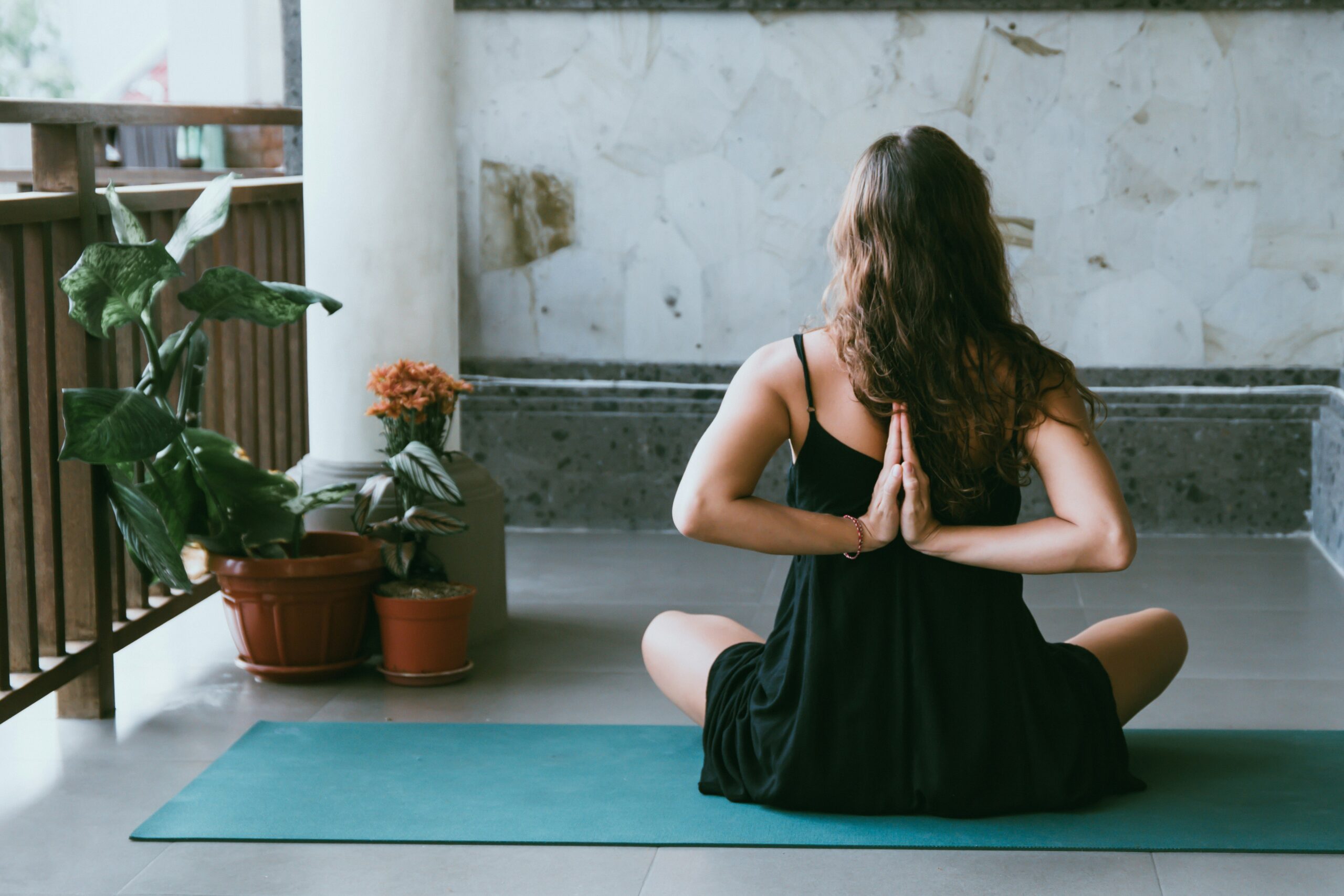 Woman practicing yoga on a mat in a peaceful indoor space, performing a reverse prayer pose, surrounded by potted plants.